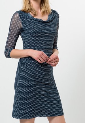 Kobieta w dopasowanej niebieskiej sukience z geometrycznym wzorem i trójczwartkowymi przezroczystymi rękawami, ręce luźno złożone na wysokości talii.