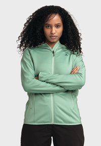 Hellgrüne Jacke mit Reißverschluss aus strukturiertem Stoff, mit Kapuze und Seitentaschen. Das Modell trägt sie mit verschränkten Armen vor einem neutralen Hintergrund.