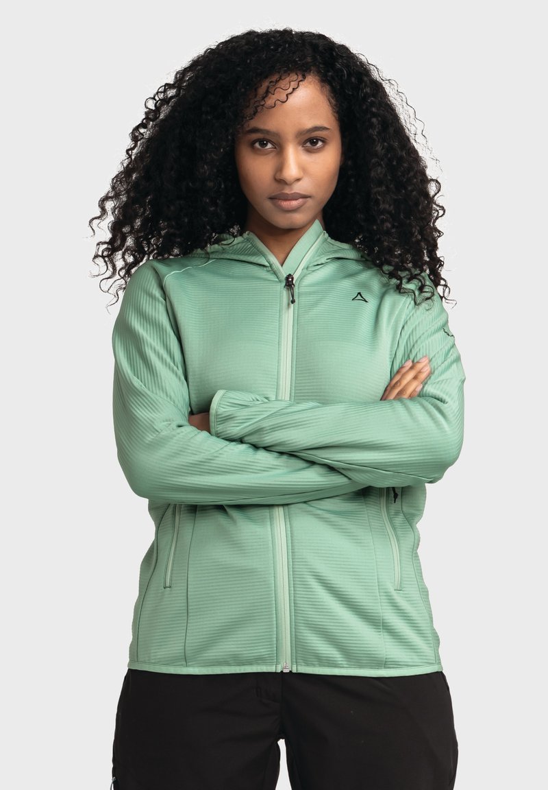 Hellgrüne Jacke mit Reißverschluss aus strukturiertem Stoff, mit Kapuze und Seitentaschen. Das Modell trägt sie mit verschränkten Armen vor einem neutralen Hintergrund.