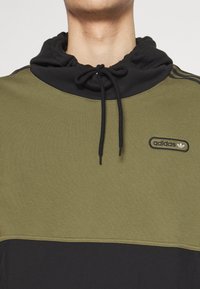 Olivgrön och svart sweatshirt med hög krage, dragsko och liten svart Adidas-logo på bröstet. Slät tygtextur.