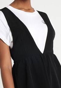 Černé šaty s hlubokým výstřihem do V přes bílé tričko. Šaty mají nařasený pas a hladkou texturu, což zvýrazňuje vrstvený vzhled.