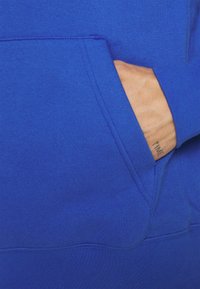 Felpa blu con tasca frontale, polsini a costine, texture liscia e dettagli di cucitura visibili.