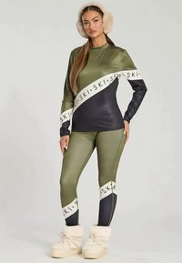 Olijfgroene en marineblauwe ski-outfit met lange mouwen en nauwsluitende legging, voorzien van logo-accenten en een gladde, glanzende textuur. Laarzen met bontvoering.