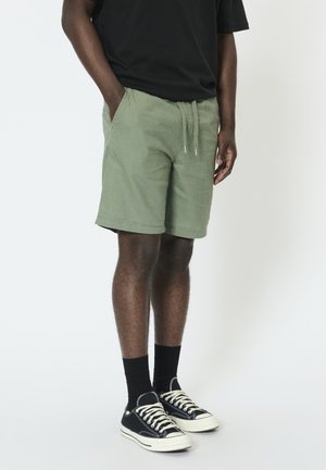 Mand iført olivengrønne shorts med snøre, sort t-shirt, sorte sokker og sorte lave sneakers, stående med hænderne i lommerne.