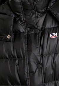 Chaqueta de abrigo negra con una textura brillante y acolchada, que presenta botones a presión negros y un parche con el logo de Levi's en la parte inferior derecha.