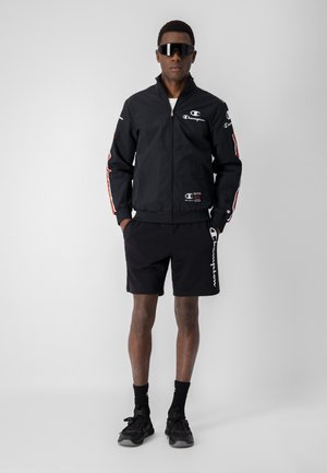 Chaqueta ligera negra con cremallera en el frente y rayas rojas en las mangas, combinada con pantalones cortos negros que tienen un logo blanco en el lado. Zapatillas negras.