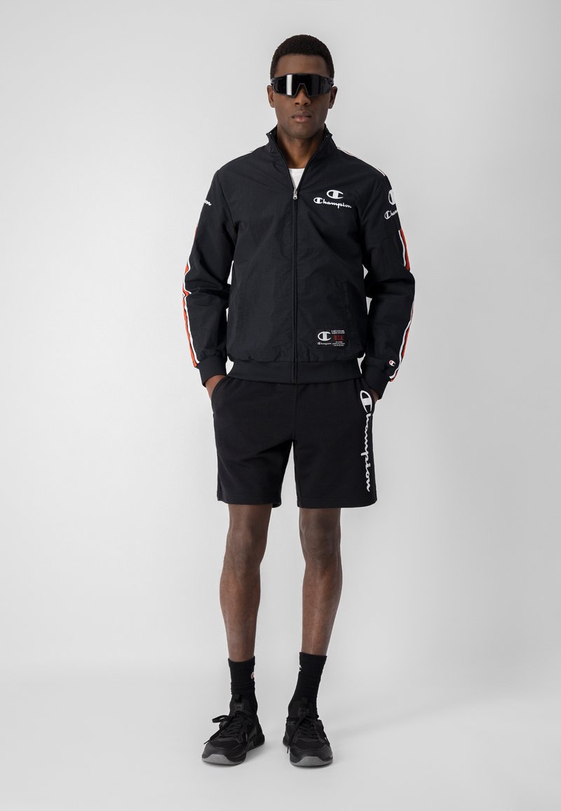 Giacca leggera nera con zip frontale e strisce rosse sulle maniche, abbinata a pantaloni corti neri con un logo bianco di lato. Sneaker nere.