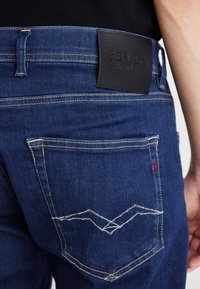 Jeans de mezclilla azul oscuro con un parche de cuero en la cinturilla, costuras intrincadas en los bolsillos traseros y un diseño ajustado.