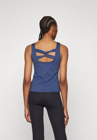 Kobieta z warkoczami, ubrana w niebieski bezrękawnik z krzyżującymi się paskami z tyłu i czarne dopasowane legginsy, stoi tyłem do aparatu.