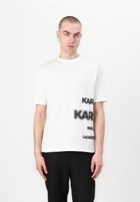 Bílá bavlněná trička s černým tečkovaným textem, který zní "KARL" a "LAGERFELD" v tučném grafickém designu. Krátké rukávy a kulatý výstřih.