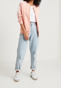 Vaaleanpunainen vetoketjullinen takki, jossa kolme valkoista raitaa hihoissa, yhdistetty vaaleansinisiin korkeavyötäröisiin farkkuihin ja valkoisiin tennareihin.