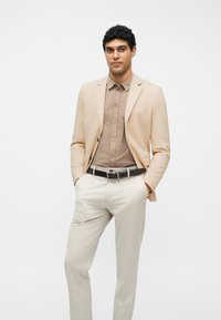 Άνδρας που στέκεται με τα χέρια στις τσέπες φοράει μπεζ σακάκι, γαλάζιο καφέ πουκάμισο με κουμπιά, λευκό παντελόνι και μαύρη ζώνη, μπροστά σε λευκό φόντο.