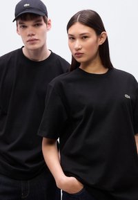 Μαύρα βαμβακερά μπλουζάκια με κοντά μανίκια, με μικρό πράσινο λογότυπο Lacoste. Ένα μοντέλο φοράει ένα μαύρο καπέλο που ταιριάζει. Ουδέτερο φόντο.