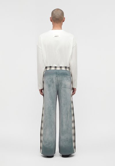 Άτομο με ξυρισμένο κεφάλι που φορά λευκό τοπ με μακριά μανίκια και φαρδιά μπλε-γκρι παντελόνια με καρό πλαϊνές ρίγες, γυρισμένο από πίσω.