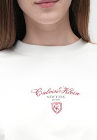 Fiatal személy fehér inget visel, amelyen piros "Calvin Klein New York EST 1968" felirat és logó hímzett a mellrészen.
