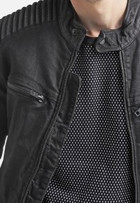 Veste en cuir noir avec épaules côtelées, poche zippée et un motif intérieur texturé de petits diamants blancs sur fond sombre.