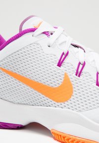 Białe buty sportowe z teksturowaną siateczką na górze, pomarańczowy znak Nike, purpurowe akcenty i białe sznurowadła; mają purpurową i pomarańczową podeszwę.