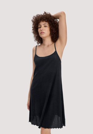 Frau mit lockigem Haar, die ein ärmelloses schwarzes Strickkleid mit dünnen Trägern trägt und mit einem erhobenen Arm vor einem schlichten Hintergrund posiert.