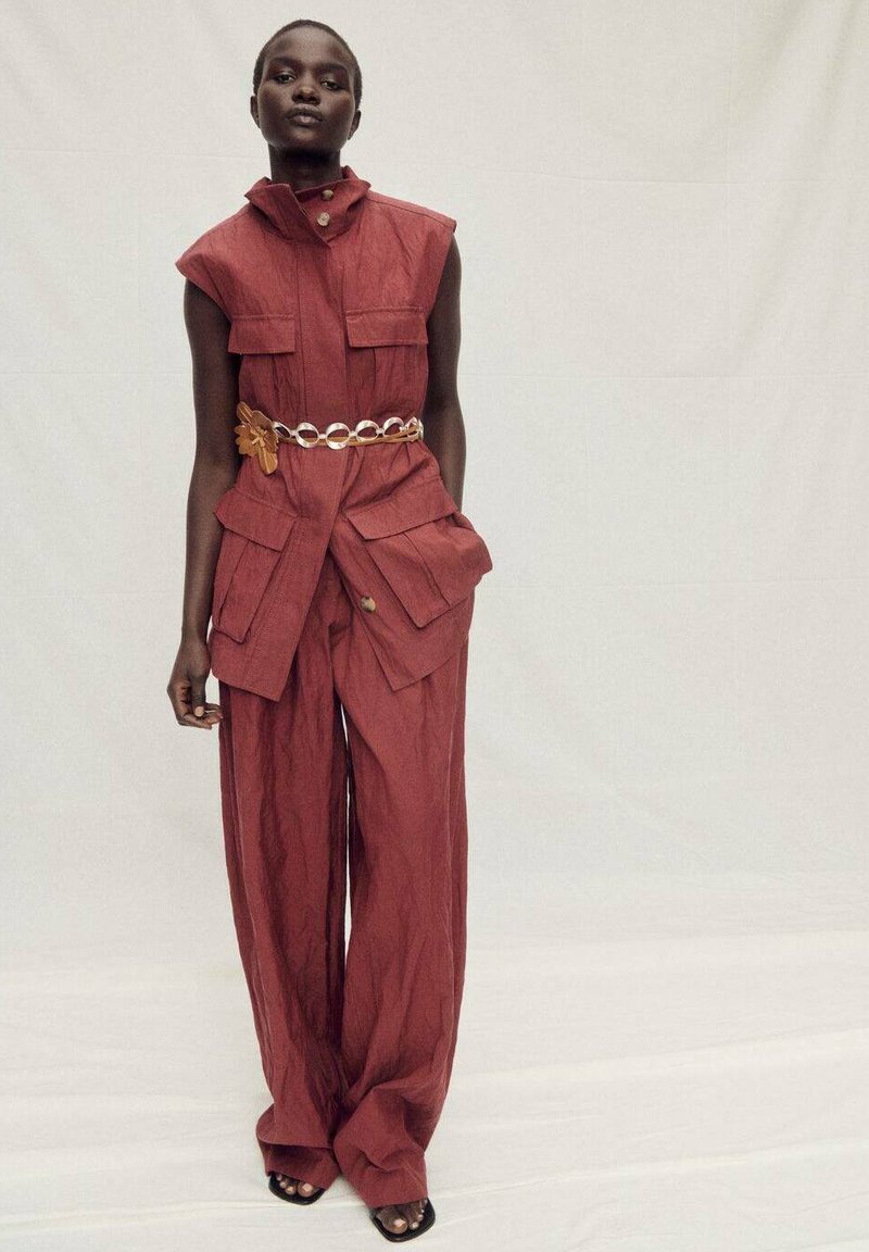 Bordeaux-rode mouwloze jumpsuit met gestructureerde kraag, voorzien van borstzakken en een wijde pijpdesign. Verfraaid met een gouden kettingriem.