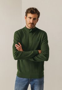 Donkergroene gebreide pullover met een hoge kraag, geribbelde manchetten en een geribbelde onderrand. Heeft een textuurstof en een klein logopatch op de borst.