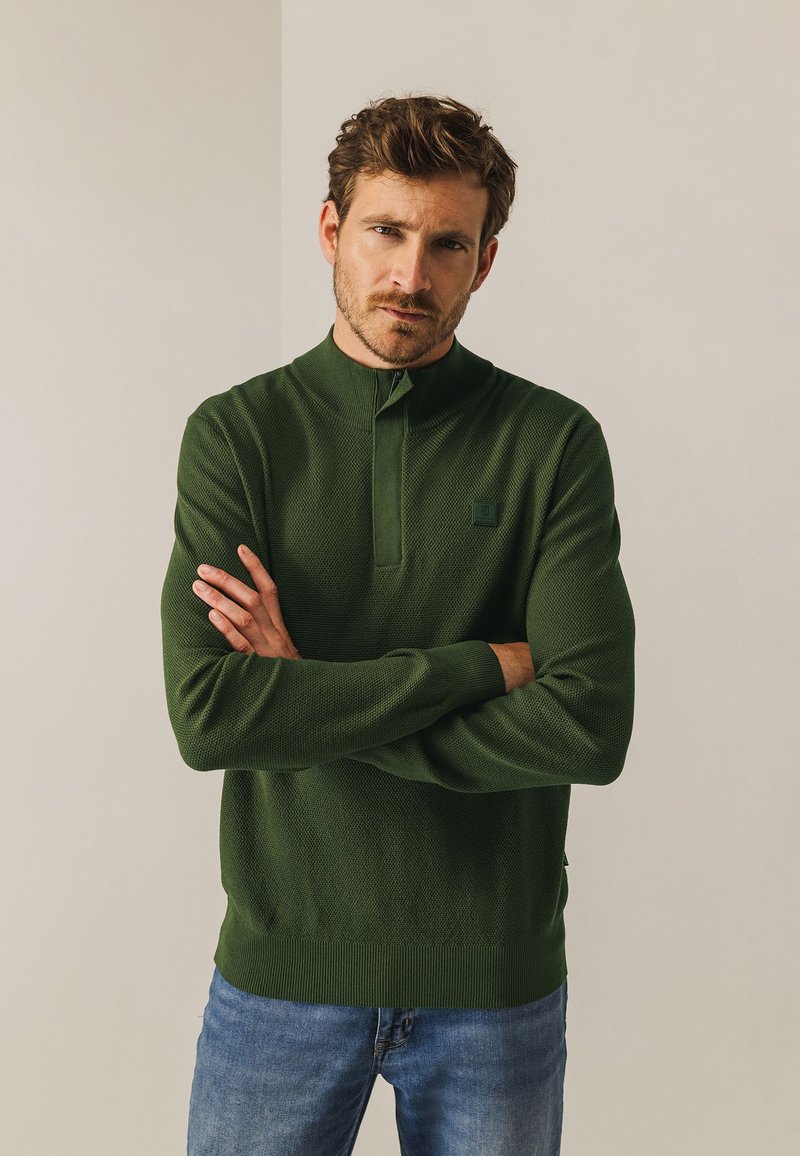 Donkergroene gebreide pullover met een hoge kraag, geribbelde manchetten en een geribbelde onderrand. Heeft een textuurstof en een klein logopatch op de borst.