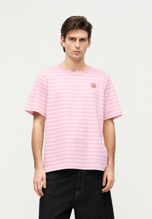 Bărbat cu păr închis la culoare poartă un tricou cu dungi orizontale roz și roz deschis cu un mic emblema cu floare roșie pe pieptul stâng, asortat cu pantaloni negri.