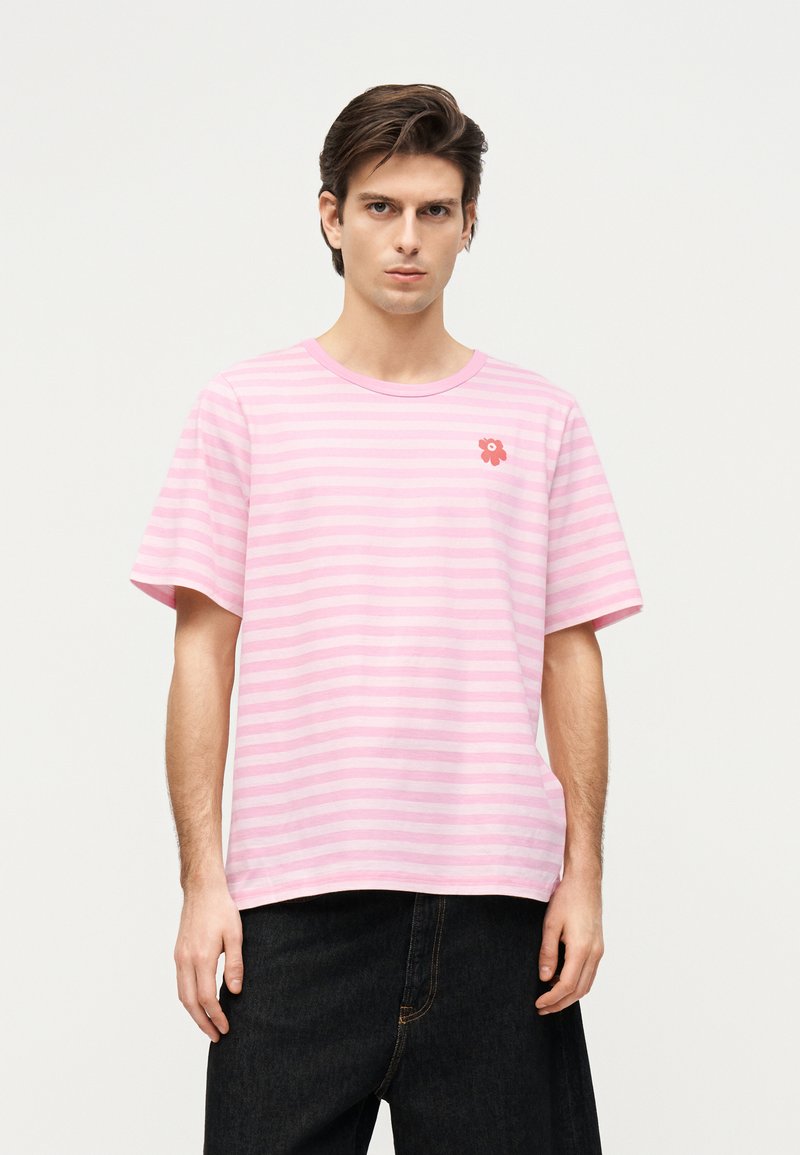 Moški s temnimi lasmi nosi majico roza in svetlo roza barve z vodoravnimi črtami ter majhen rdeč cvetlični emblem na levi strani prsnega koša, v kombinaciji s črnimi hlačami.