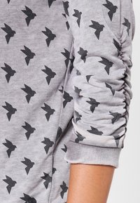 Felpa grigia con un motivo di uccelli neri. Le maniche presentano una texture arricciata e i polsini a costine aggiungono definizione.