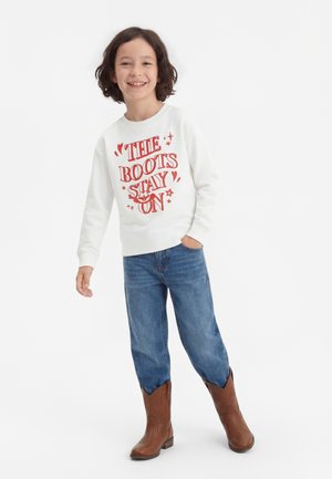 Uśmiechnięte dziecko o kręconych włosach nosi białą bluzę z napisem "The Boots Stay On", niebieskie dżinsy i brązowe kowbojki, stoi na białym tle.