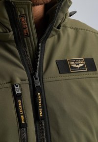 Maslinasto zelena jakna s ušivenim uzorkom, s dvije džepove na zatvarač i crnim akcentima. Logotip na prsima i zatvarači s brendiranjem.