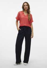 Blusa corallo a maniche corte con scollo a V; abbinata a pantaloni neri a vita alta con cintura a fiocco. Tessuto morbido, vestibilità rilassata.