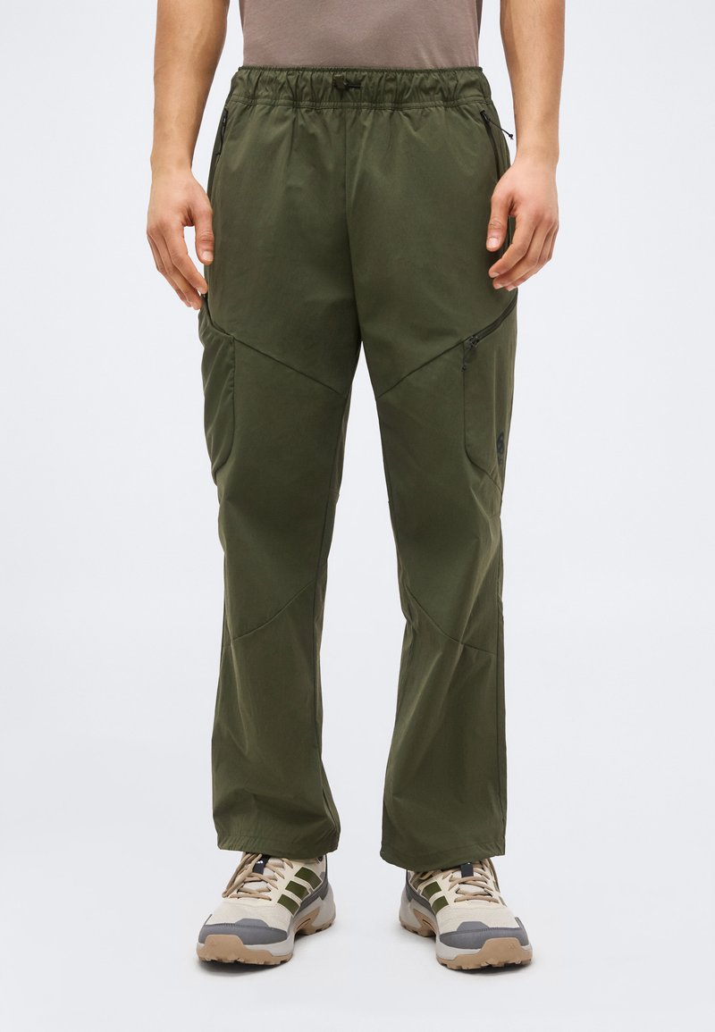 Férfi, aki olíva zöld szabadtéri nadrágot visel oldalzsebes cipzárral, valamint bézs túracipőt zöld és szürke részletekkel.