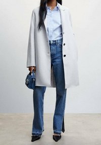 Lichtgrijze oversized jas, blauwe shirt, hoge taille blauwe jeans met zijsplitten en zwarte puntige hakken. Kleine blauwe handtas.