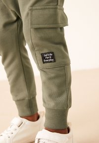 Maslinasto zelene trenirka hlače s džepovima na boku, rebrastim manžetama i crnom oznakom s bijelim tekstom. Uparene s bijelim tenisicama.
