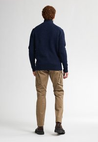 Pull en col roulé tricoté bleu marine avec un tissu texturé, complété par un pantalon cargo beige. Des chaussures foncées viennent parfaire la tenue.