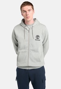 Ung mand med kort brunt hår iført en grå Timberland hættejakke med lynlås og marineblå cargo-bukser, stående med hænderne bag ryggen.