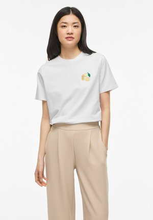 Nő fehér pólót visel, melyen apró hímzett citrom minta található, bézs színű, bőszárú nadrágban áll egy egyszerű háttér előtt.