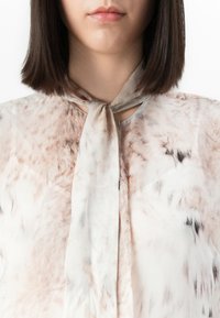 Frau mit glattem, dunklem Haar, die eine hellmusterte Bluse mit einem gebundenen Halstuch trägt, vom Brust- bis Lippenbereich vor einem weißen Hintergrund gezeigt.