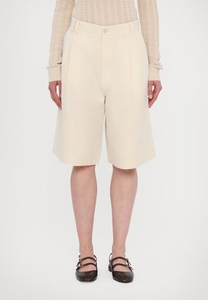 Short beige sur mesure longueur genou porté avec un haut léger en maille à manches longues et des chaussures plates marron foncé à double bride.