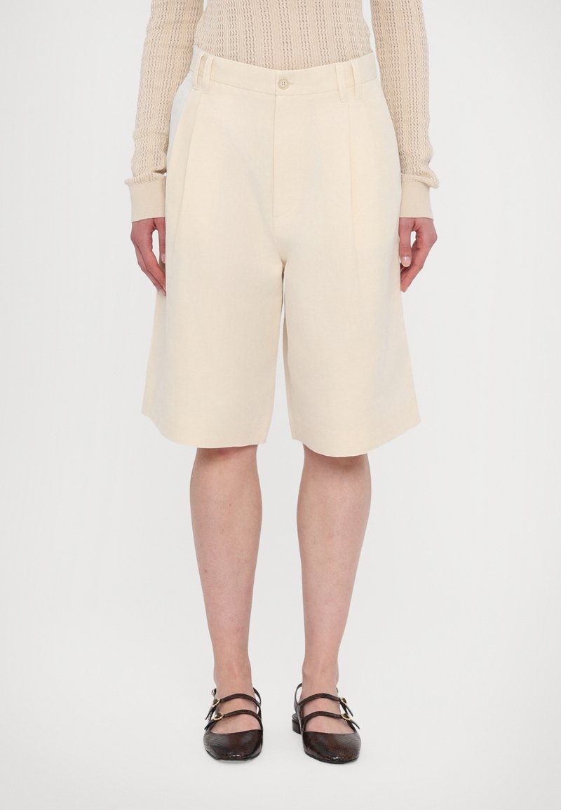 Short beige sur mesure longueur genou porté avec un haut léger en maille à manches longues et des chaussures plates marron foncé à double bride.