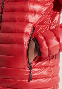 Rote Steppjacke mit glänzendem Finish, horizontalem Steppmuster und einer Reißverschlusstasche. Der Ärmel hat eine Rippbündchen mit kontrastierendem Stoff.