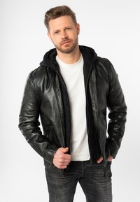 Giubbotto in pelle nera con cappuccio, apertura con zip e due tasche laterali. Indossato sopra una maglietta bianca e jeans grigi, con un interno testurizzato.