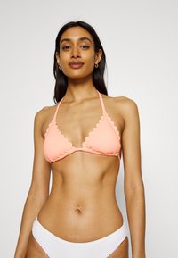 Top de bikini cor-de-rosa coral com copas triangulares, bordas em forma de concha e alças ajustáveis. Combinado com parte de baixo de bikini branca texturizada.