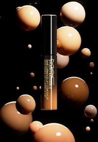 El corrector luminoso de MAC Studio Radiance 24HR tiene un elegante tubo negro con letras doradas, rodeado de varias tonalidades de esferas beige brillantes.