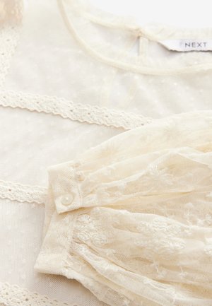 Bluză din dantelă în crem, cu broderie florală, material transparent și închidere cu buton. Include o margine delicată de-a lungul gâtului și manșetelor.