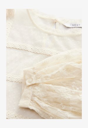 Blouse en dentelle crème, ornée de broderies florales, en tissu transparent, avec une fermeture à boutons. Inclut un délicat contour le long de l'encolure et des manches.