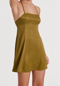 Robe sans manches vert olive en tissu lisse et satiné, avec un corsage ajusté et une jupe évasée descendant au-dessus du genou.