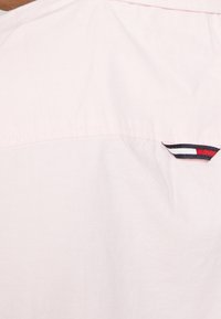 Jasnoróżowa koszula z teksturowanej tkaniny, z kołnierzykiem na guziki oraz małą naszywką z logo i czerwonymi, białymi i niebieskimi akcentami na piersi.