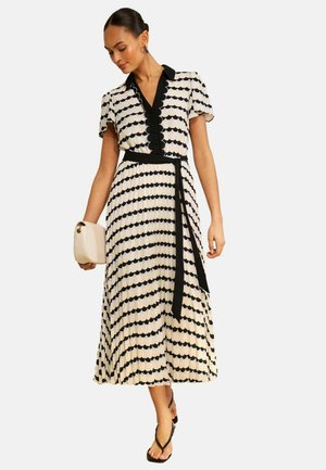 Nő fekete-fehér hullámos szegélyű midi ruhában, fekete övvel, bézs táskával a kezében, fekete szandált és kék fülbevalót visel.