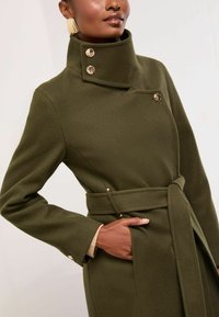 Olivroheline villane mantel kõrge kraega, kuldsete nööpidega ja vöödiga taljes. Omab struktureeritud õlgu ja sileda, tekstureeritud viimistlust.